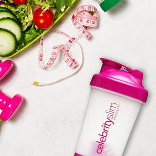 Overhead view of a Celebrity Slim blender with a measuring tape, pink dumbbells, and fresh salad ingredients on a green tray.