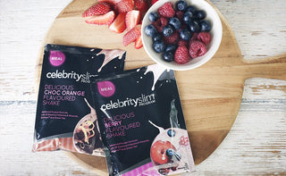 Overhead shot of Celebrity Slim shake sachets on a wooden plate, with sliced strawberries and a bowl of assorted berries.