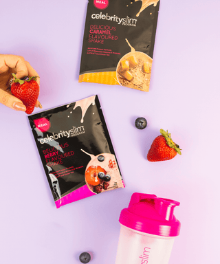 High Angle Shot: A person holds a strawberry, surrounded by two Celebrity Slim sachets , a blender, and berries.